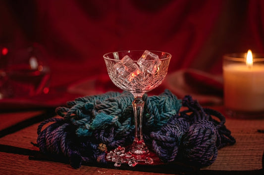 Turquoise and purple silk bondage ropes next to ice in a crystal glass and a candle flame in a dark, red-lit mood.