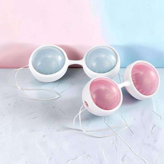 Two pairs of white silicone strap-on toys with translucent domes, one pair in light blue and the other in pink, arranged on a pastel pink and blue background.