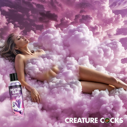 Woman lying on pink clouds with a bottle of 'Creature Cocks Creature Slime - Purple H2O Lube 4oz' in front, against a pink sky.